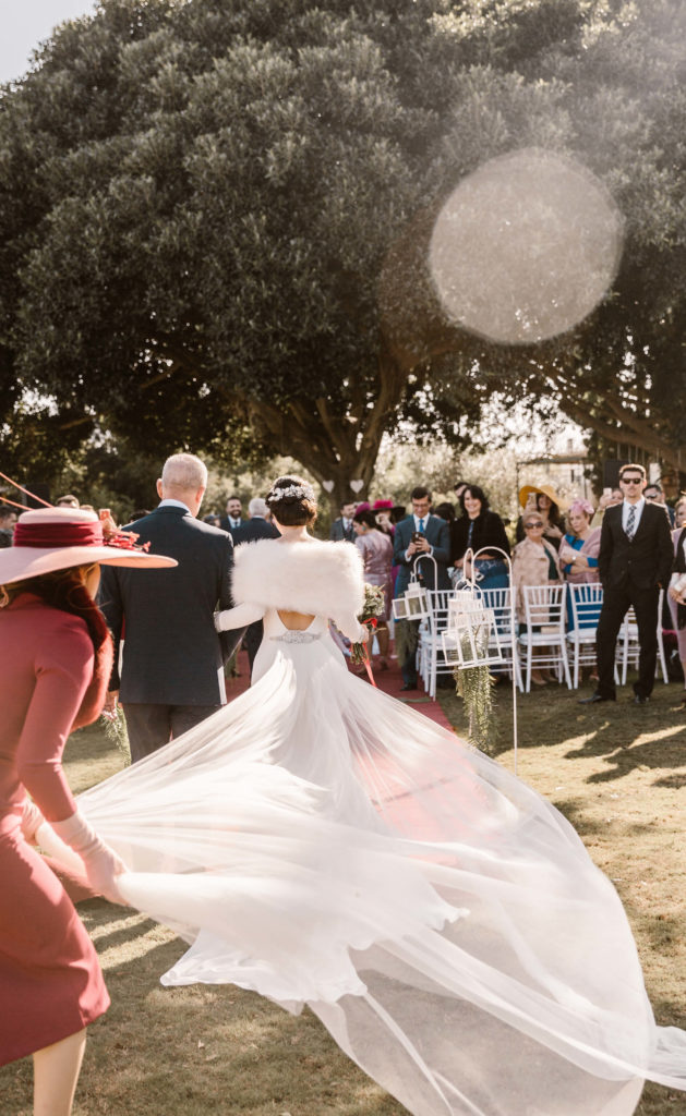 Novia caminando hacia el altar con una larga cola, vestido de manga larga para una boda en otoño en el real club de golf de Sevilla y organizada por la wedding planner flory ferreras