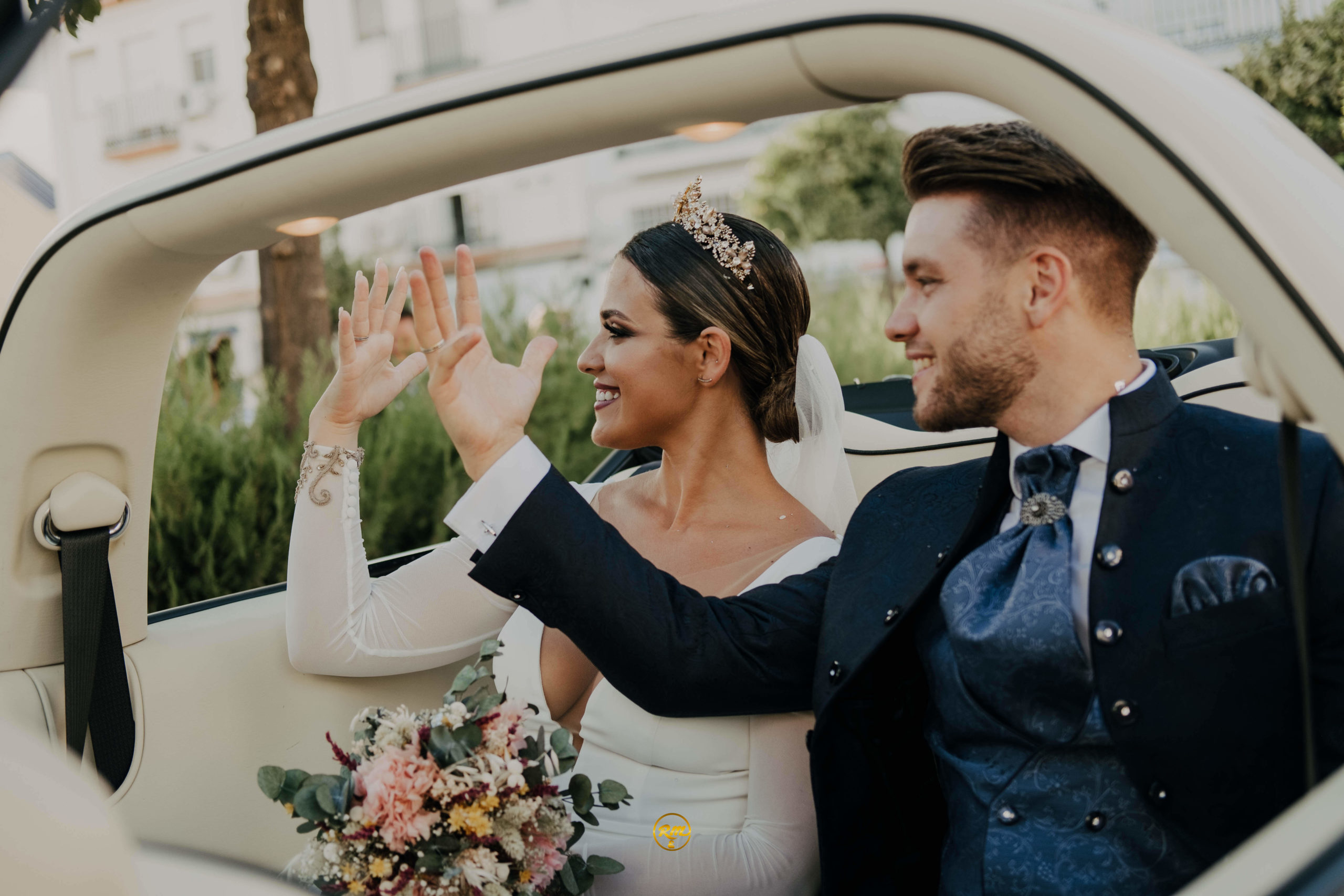 novios saludando desde el coche y enseñando los anillos tras su boda. el coche es blanco por dentro y por fuera y descapotable