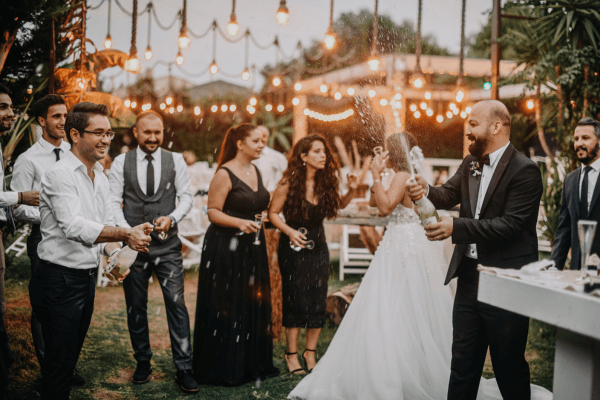 novio agitando el champán en una boda, con los invitados y luces decorativas al fondo